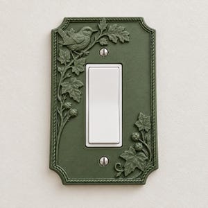 May include: A rectangular olive green light switch cover plate featuring a raised relief design of a bird perched on a branch with leaves and berries. The plate has a rope-style border, a central white rocker switch, and two silver screws.