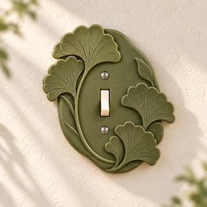 May include: An olive green light switch plate featuring a raised, sculpted ginkgo leaf design. The plate has an oval shape with a central vertical toggle switch in cream and two silver-toned screws securing the cover to the wall.