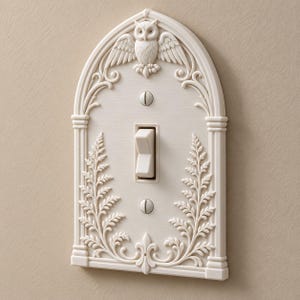 May include: An off-white light switch cover plate featuring an arched top with a sculpted owl at the peak and symmetrical fern leaf carvings along the sides. The rectangular plate includes a central toggle switch and two visible mounting screws.