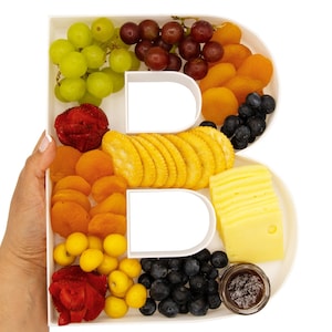 May include: A white ceramic letter B shaped serving tray filled with various snacks, including grapes, strawberries, dried apricots, crackers, cheese, blueberries, and a small jar of jam.