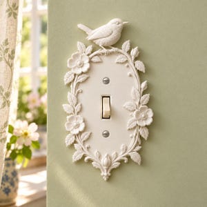 May include: An off-white light switch plate featuring a sculpted relief of a bird perched on a branch at the top, surrounded by a decorative border of embossed flowers, leaves, and buds. The plate includes two silver screws and a toggle switch.