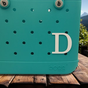 May include: Teal blue Bogg bag with a white letter D on the front. The bag has a textured surface with small holes.