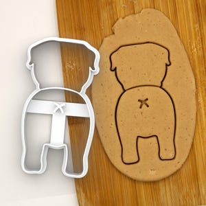 May include: A white plastic cookie cutter shaped like a dog's rear end, with a cross on the tail, is shown next to a brown cookie dough cutout of the same shape.