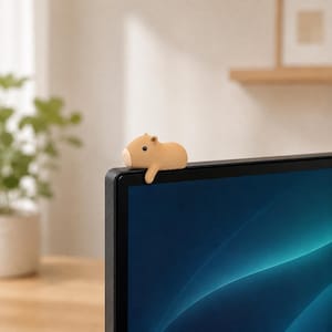 May include: A small, tan, capybara-shaped figure with a rounded body, tiny ears, and a dark eye, perched on the top edge of a black computer monitor. The figure has a smooth, matte texture and is positioned to look as if it is resting on the screen frame.
