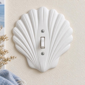 May include: A white light switch cover plate shaped like a scallop shell. The cover features a central vertical toggle switch and is secured to the wall with two visible silver-toned metal screws positioned above and below the switch toggle.