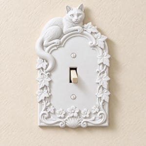 May include: A white light switch plate featuring a sculpted cat resting on the top edge, surrounded by embossed ivy leaves and floral details. The rectangular cover includes a central toggle switch and two visible mounting screws.