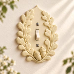 May include: A cream-colored light switch plate featuring a raised relief design of oak leaves framing the central toggle switch. Two small acorns are positioned at the bottom, and the plate is secured with two visible silver-toned metal screws.