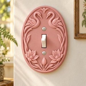 May include: An oval-shaped, dusty pink light switch cover plate featuring a raised relief design of two swans facing each other at the top and two blooming lotus flowers at the bottom. The plate includes a white toggle switch and two silver screws.