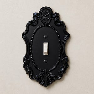 May include: A black light switch plate with an ornate, oval-shaped frame featuring decorative scrollwork and a beaded border. The plate includes a single white toggle switch in the center, secured by two black screws positioned above and below.