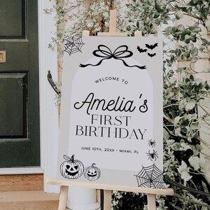 Puede incluir: Un caballete de madera muestra un letrero para el primer cumpleaños de Amelia. El letrero tiene un fondo claro con ilustraciones negras con temática de Halloween, que incluyen calabazas, murciélagos y telarañas. El texto dice "Welcome to Amelia's First Birthday."