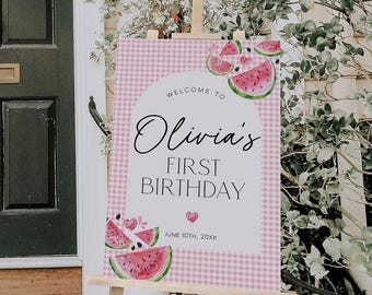 Puede incluir: Un letrero rectangular de bienvenida de cumpleaños sobre un caballete de madera con un patrón de cuadros vichy rosa y blanco, rodajas de sandía en acuarela y el texto: Welcome to Olivia's First Birthday, June 10th, 20XX, con arco central.