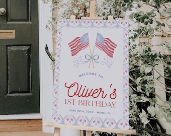 May include: A rectangular welcome sign on a wooden easel featuring two crossed American flags with a blue bow, red and blue fireworks, and text reading: Welcome to Oliver's 1st Birthday, June 10th, 20XX, Miami, FL, framed by a red and blue patterned border.