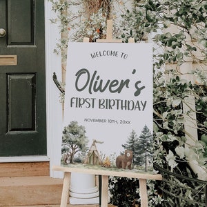 May include: Welcome to Oliver's First Birthday sign with a woodland theme. The sign features a white background with black text and a watercolor illustration of a forest scene with a tent, trees, a bear, and a squirrel.
