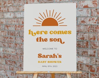 May include: A rectangular white sign on a wooden easel featuring an orange sun graphic and the text: Here comes the son, WELCOME TO Sarah's BABY SHOWER APRIL 10TH, 2023. The sign uses orange and yellow lettering in a retro-inspired font style.