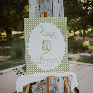 May include: A wooden easel supports a sign for a bridal shower. The sign has the text "The Perfect Pear" with pear illustrations and event details. The sign is white with green accents and a decorative border.