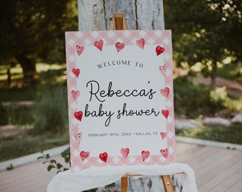 May include: A rectangular sign with a pink and white gingham border featuring red heart illustrations. The text reads: WELCOME TO Rebecca's baby shower FEBRUARY 10TH, 20XX • DALLAS, TX. The sign is displayed on a wooden easel with a white cloth draped over it.