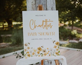 May include: A white rectangular sign on a wooden easel featuring the text WELCOME TO Charlotte's BABY SHOWER JUNE 10TH, 20XX DALLAS, TX in gold and black lettering, decorated with a watercolor illustration of yellow flowers and small bees.