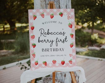 May include: A rectangular welcome sign with a pink and white gingham border decorated with strawberry illustrations. The text reads: WELCOME TO Rebecca's berry first BIRTHDAY JUNE 10TH, 20XX • DALLAS, TX. The sign is displayed on a wooden easel.
