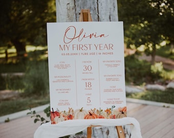 May include: A white milestone sign titled Olivia My First Year with birth details 7lbs 2oz and 19 inches. The board lists developmental milestones, personality traits, and favorites, decorated with a colorful watercolor pumpkin and floral border.