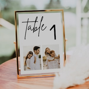 May include: A gold-framed table number sign with the text "Table 1" in black script font. Two black and white photos are displayed below the text, showing a couple in different poses.