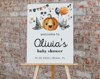 May include: A white rectangular sign featuring a Halloween-themed illustration of a pumpkin, a witch hat, a bat, and balloons. The text reads: welcome to Olivia's baby shower 10.30.2023 | Miami, FL. The sign is displayed on a wooden easel.