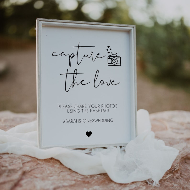 May include: White sign with black text that reads "capture the love".  A small black and white camera icon is above the text.  Below the text is "Please share your photos using the hashtag! #SARAH&JONESWEDDING"
