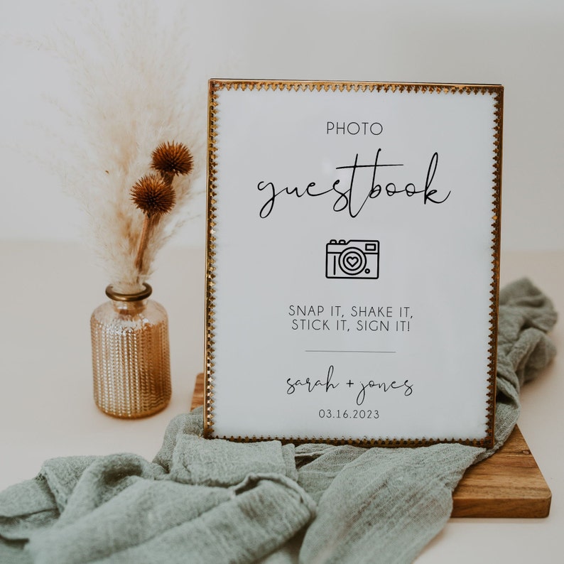 May include: A gold framed sign with a white background and black text that reads "PHOTO guestbook" with a camera icon and the text "SNAP IT, SHAKE IT, STICK IT, SIGN ITI" and "sarah + jones 03.16.2023".