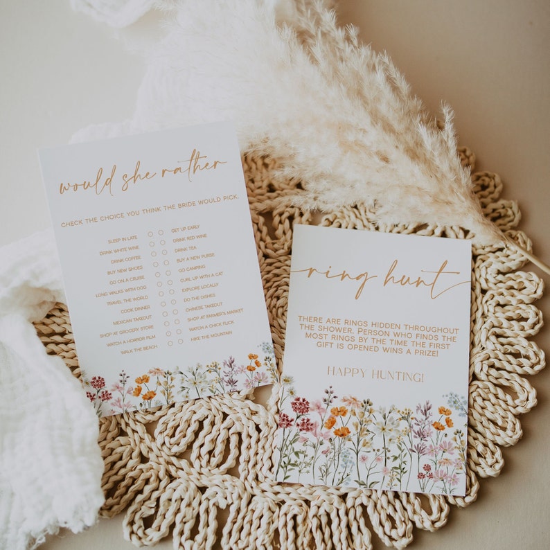 May include: Two white cards with gold text and floral borders. One card reads "Would She Rather" with a list of choices. The other says "Ring Hunt" with instructions for a game. Both cards are on a woven mat.