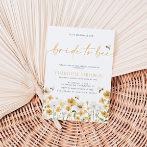 May include: A white bridal shower invitation with gold lettering that says "Let's Celebrate the Bride to Bee". The invitation has a floral design with yellow and white flowers and bees.