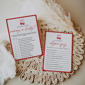 May include: Two baby shower game cards with a red cherry design. One card reads "Guess Who mommy or daddy?" and the other reads "Nursery Rhyme Quiz." Both cards have a scalloped border and are on a cream-colored, woven surface.