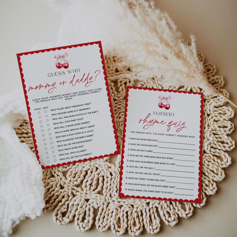 May include: Two baby shower game cards with a red cherry design. One card reads "Guess Who mommy or daddy?" and the other reads "Nursery Rhyme Quiz." Both cards have a scalloped border and are on a cream-colored, woven surface.