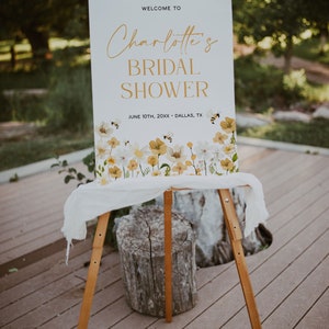 K&ouml;nnte beinhalten: Ein wei&szlig;es Schild mit goldenen Buchstaben, das "Willkommen zu Charlotte's Bridal Shower" mit dem Datum "10. Juni 20XX - Dallas, TX" ank&uuml;ndigt. Das Schild ist mit einer Aquarell-Illustration von gelben und wei&szlig;en Blumen und Bienen verziert.