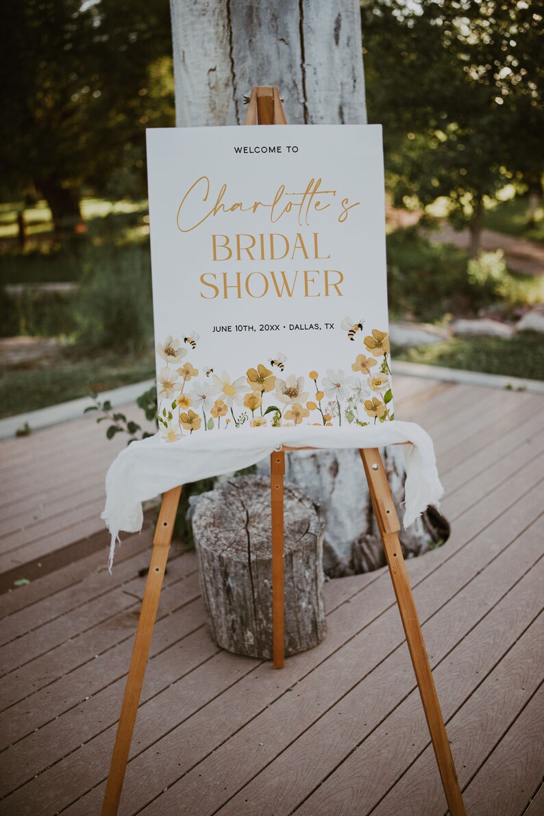 K&ouml;nnte beinhalten: Ein wei&szlig;es Schild mit goldenen Buchstaben, das "Willkommen zu Charlotte's Bridal Shower" mit dem Datum "10. Juni 20XX - Dallas, TX" ank&uuml;ndigt. Das Schild ist mit einer Aquarell-Illustration von gelben und wei&szlig;en Blumen und Bienen verziert.