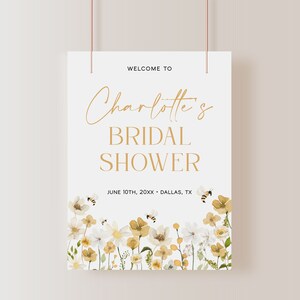 K&ouml;nnte beinhalten: Ein wei&szlig;es Brautdusche-Schild mit goldenen Buchstaben, auf dem "Willkommen zu Charlotte's Bridal Shower" und "June 10th, 20XX - Dallas, TX" steht. Das Schild ist mit einem Aquarell-Blumenrand aus wei&szlig;en und gelben Blumen und Bienen verziert.