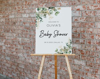 May include: A rectangular white sign on a wooden easel featuring green eucalyptus leaf illustrations in the top left and bottom right corners. The text reads: WELCOME TO OLIVIA'S Baby Shower 08.10.2023 | DALLAS, TX in black lettering.