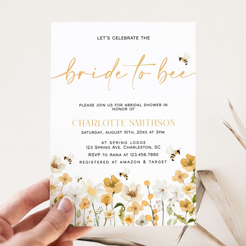 May include: A white bridal shower invitation with a gold script font that says "bride to bee". The invitation has a floral border with yellow and white flowers and bees. The text on the invitation reads "Let's celebrate the bride to bee. Please join us for a bridal shower in honour of Charlotte Smithson. Saturday, August 10th, 20XX at 3PM. At Spring Lodge, 123 Spring Ave. Charleston, SC. RSVP to Anna at 123.456.7890. Registered at Amazon & Target."