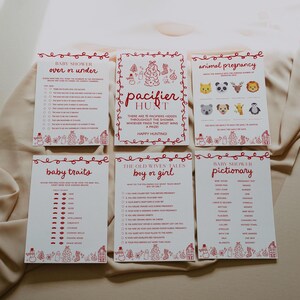 May include: Six baby shower game cards with red text and illustrations on a cream background. Games include "Over or Under," "Pacifier Hunt," "Animal Pregnancy," "Baby Traits," "Boy or Girl," and "Baby Shower Pictionary."