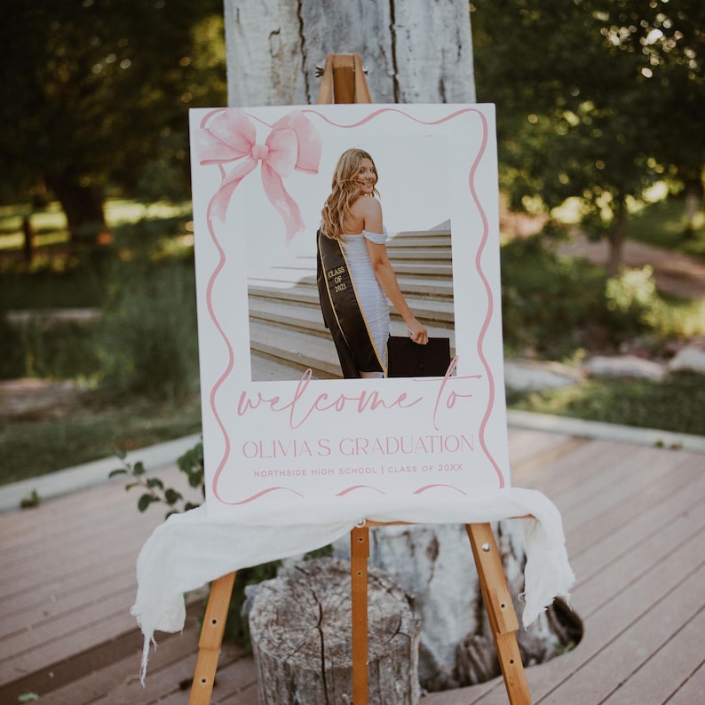 May include: A white welcome sign with pink lettering and a pink bow. The sign says "Welcome to Olivia's Graduation, Northside High School, Class of 20XX". A photo of a graduate in a cap and gown is in the center of the sign.