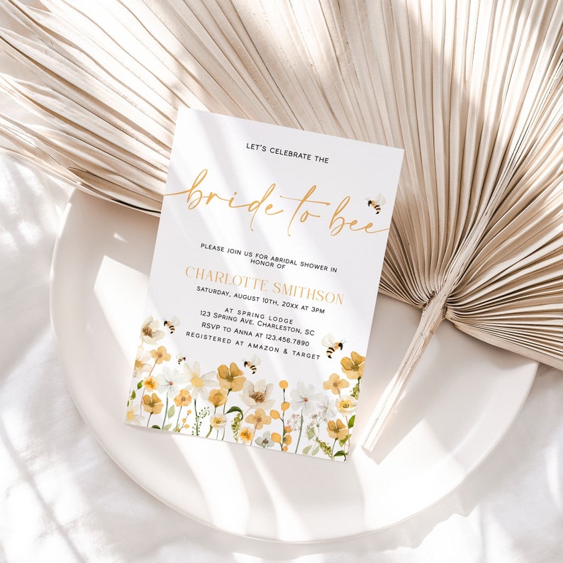 May include: A white bridal shower invitation with gold lettering that says "Let's Celebrate the Bride to Bee". The invitation has a floral design with yellow and white flowers and bees.