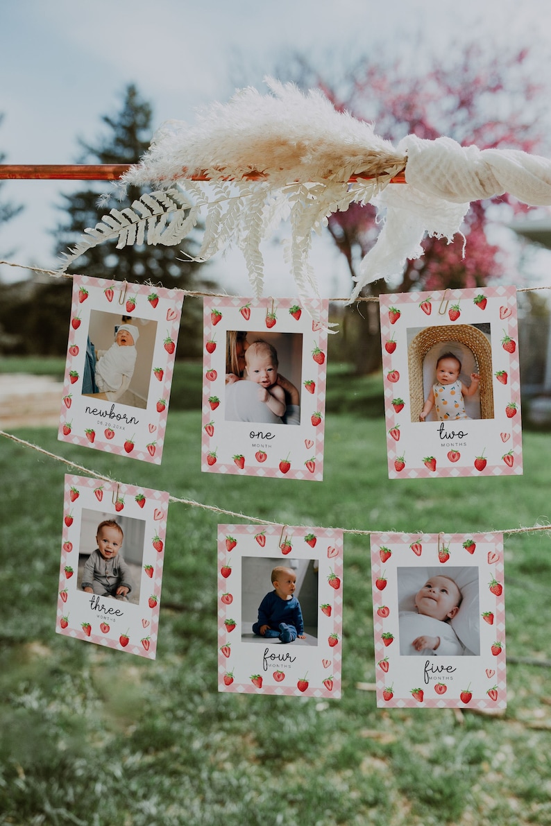 May include: A set of six pink and white photo cards with strawberry designs. Each card features a photo of a baby at different ages, with the age in months written below the photo. The cards are strung together with twine and are perfect for a baby's first year photo shoot.