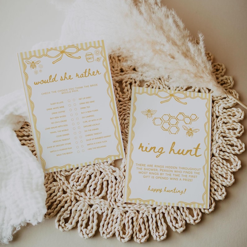 May include: Two ivory-colored cards with gold accents and text for a bridal shower game. One card says "Would She Rather" with a list of choices. The other card says "Ring Hunt" with bee and honeycomb graphics.