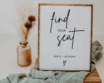 May include: A gold-framed sign with a white background and black text that reads "find your seat" with the names "Grace + Anthony" and a heart symbol below. The sign is sitting on a wooden surface with a green cloth draped over it.