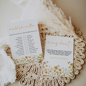 May include: Two white cards with gold text and floral accents. One card reads "Would She Rather" with a list of choices. The other card says "Ring Hunt" with instructions for a game. Both cards have a yellow and white floral design.