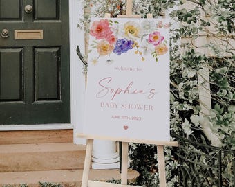 May include: A rectangular white sign on a wooden easel featuring a watercolor floral arrangement in pink, yellow, and purple at the top. The text reads: welcome to Sophia's BABY SHOWER JUNE 10TH, 2023, with a small pink heart icon below the date.