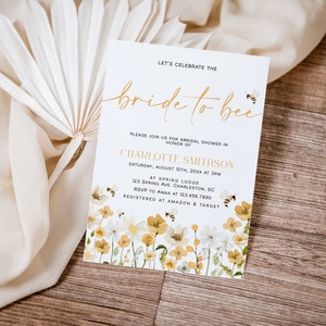 May include: A white bridal shower invitation with a gold script font that says "bride to bee". The invitation has a floral design with yellow and white flowers and bees. The text on the invitation reads "Let's celebrate the bride to bee. Please join us for bridal shower in honour of Charlotte Smithson. Saturday, August 10th, 20XX at 3PM. At Spring Lodge, 123 Spring Ave. Charleston, SC. RSVP to Anna at 123.456.7890. Registered at Amazon & Target."