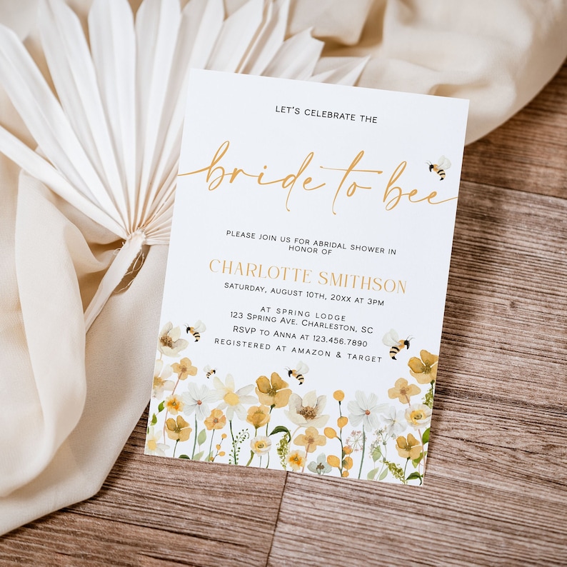 May include: A white bridal shower invitation with a gold script font that says "bride to bee". The invitation has a floral design with yellow and white flowers and bees. The text on the invitation reads "Let's celebrate the bride to bee. Please join us for bridal shower in honour of Charlotte Smithson. Saturday, August 10th, 20XX at 3PM. At Spring Lodge, 123 Spring Ave. Charleston, SC. RSVP to Anna at 123.456.7890. Registered at Amazon & Target."