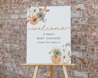May include: A rectangular white sign featuring watercolor illustrations of pumpkins and autumn leaves in the top left and bottom right corners. The text reads: welcome to Hazel's BABY SHOWER october 10th | dallas, tx in brown and black font.