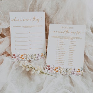 May include: Two white cards with gold text and floral accents. One card reads "where were they?" with fill-in-the-blank prompts. The other says "I love you around the world" with a matching game. A sprig of white flowers rests below.