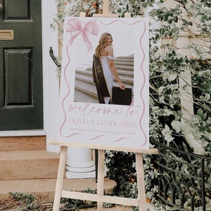May include: A white welcome sign with pink accents and a photo of a graduate in a cap and gown. The sign reads "Welcome to Olivia's Graduation, Northside High School, Class of 20XX".
