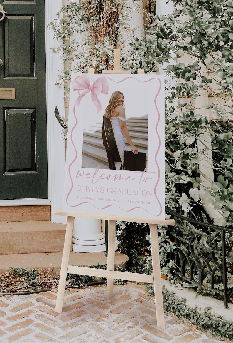 May include: A white welcome sign with pink accents and a photo of a graduate in a cap and gown. The sign reads "Welcome to Olivia's Graduation, Northside High School, Class of 20XX".